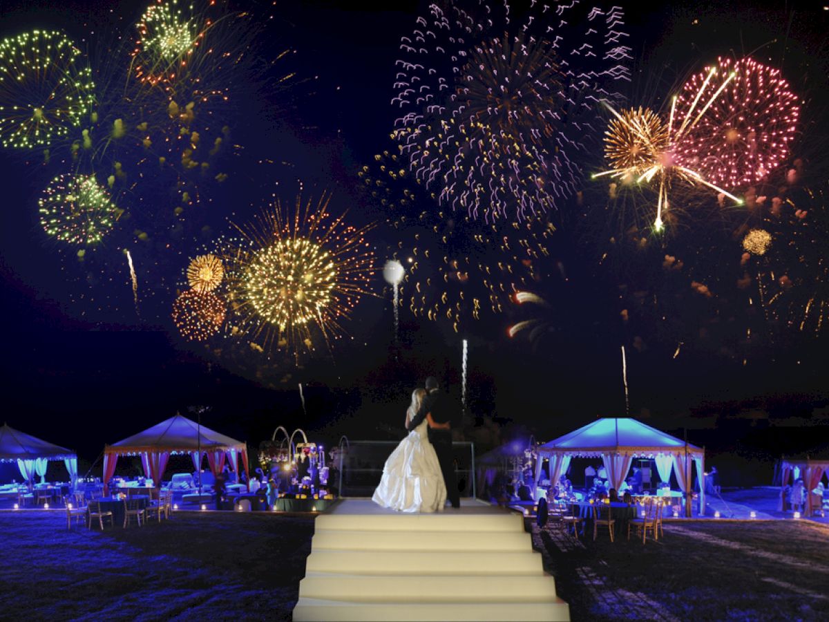 A person in a white dress stands on a staircase outdoors at night, with colorful fireworks lighting up the sky and tents lit below, ending the sentence.