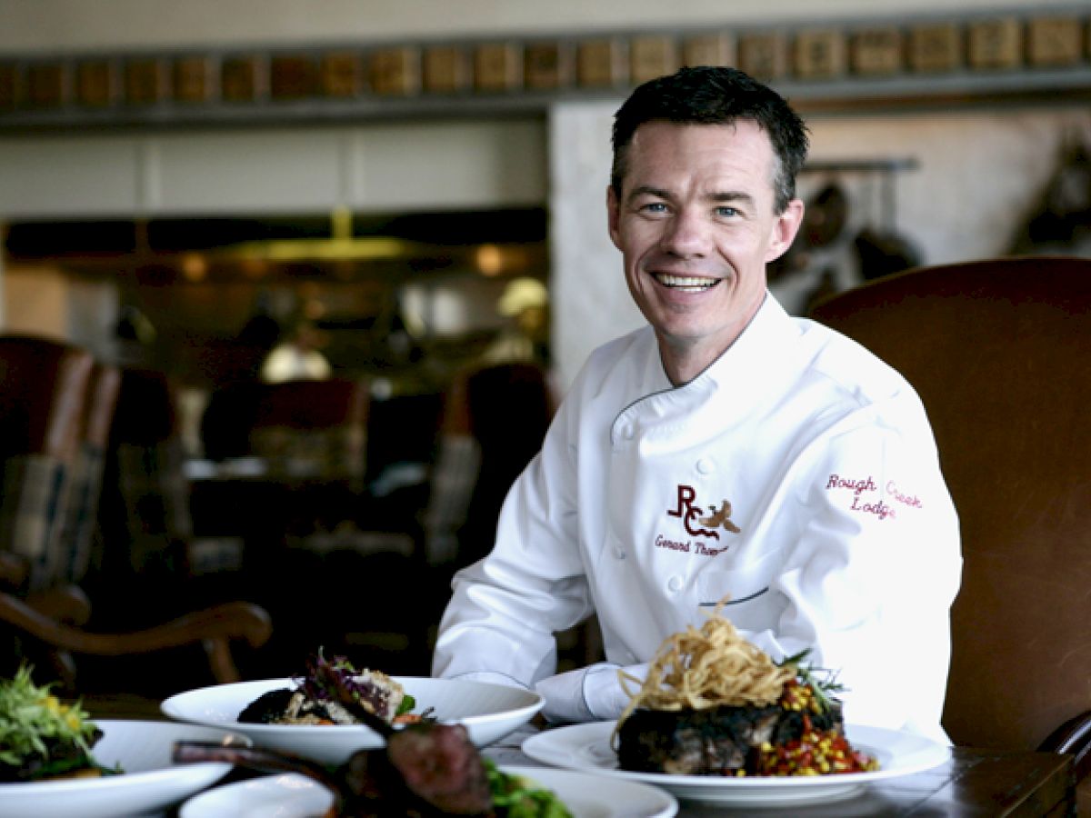 A chef in a white uniform sits at a table with several plated dishes, smiling at the camera in a restaurant setting.