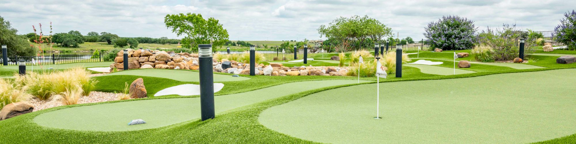 The image shows a landscaped mini-golf course with artificial turf, flags, rocks, and surrounded by greenery under a cloudy sky.