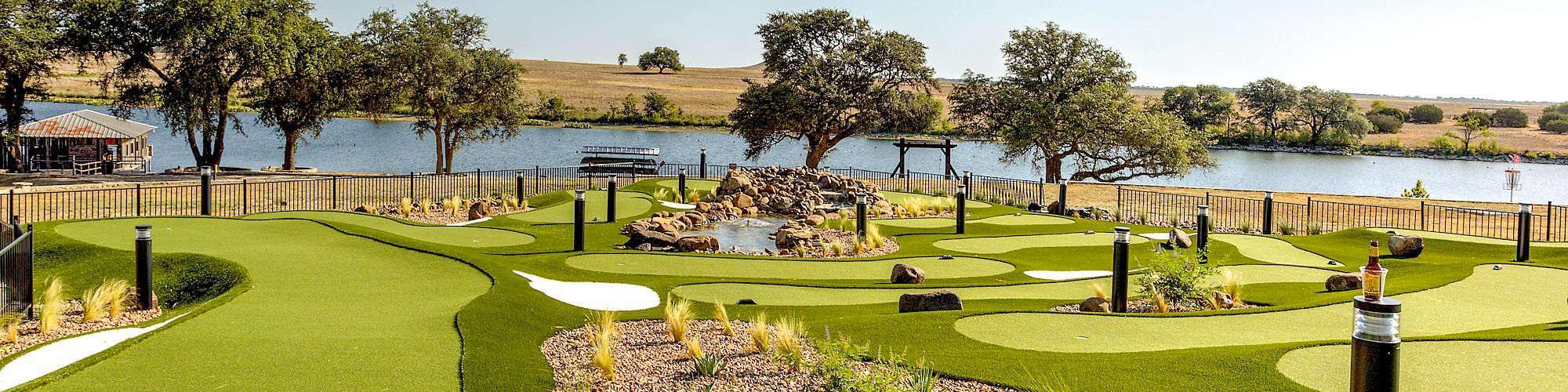 A beautifully landscaped mini-golf course by a serene lake, featuring lush green artificial turf, landscaped rock formations, and strategically placed shrubs.