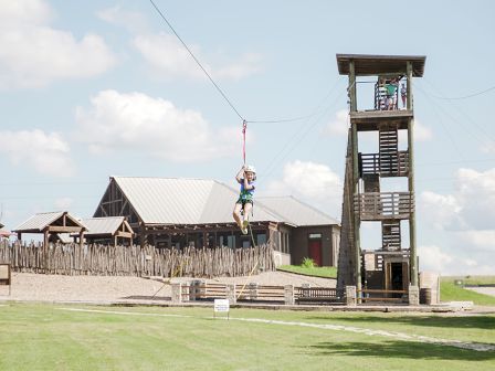 A person is zip-lining from a tall wooden tower with buildings and a fence in the background, under a clear sky with scattered clouds.