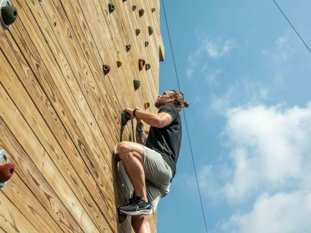 A person is climbing an outdoor wooden rock climbing wall on a sunny day. A sign at the bottom reads: "HAVE FUN THINK SAFETY."