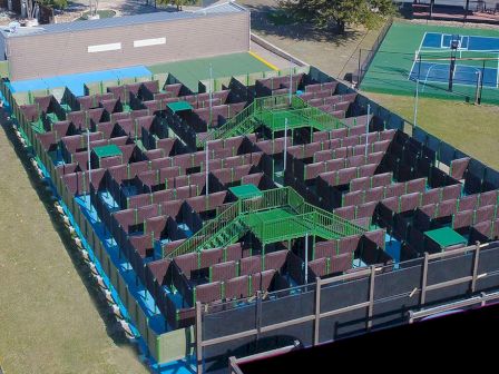 This image features an outdoor paintball maze with various pathways and barriers, situated next to a basketball court.