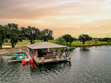 A lakeside dock with a covered boat shed and kayaks, people standing nearby as sunset colors the sky over green park grounds.