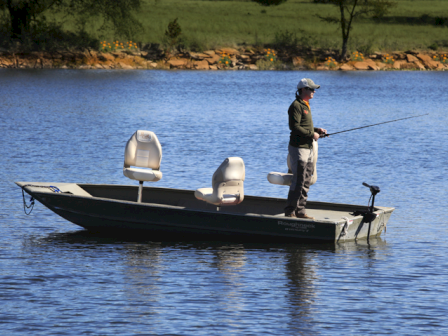 A person is fishing on a small boat in a lake with two white seats, surrounded by water and a distant shoreline.