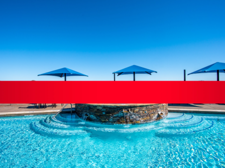 Outdoor swimming pool with clear blue water, red barrier wall, and three blue umbrellas shading lounge chairs by a stone wall.