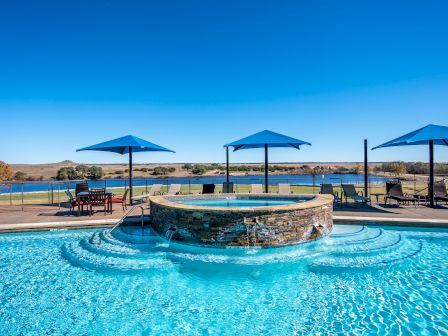 A clear pool with a circular hot tub, surrounded by blue umbrellas and loungers, overlooking a serene lake and open landscape under a blue sky.