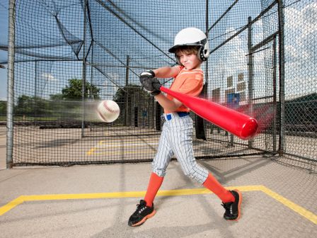 A young baseball player in a batting cage swings a red bat at a baseball, wearing an orange shirt, helmet, and striped pants.