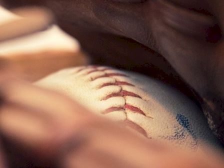 A close-up of a baseball being held by a hand is shown, highlighting the red stitching on the ball.