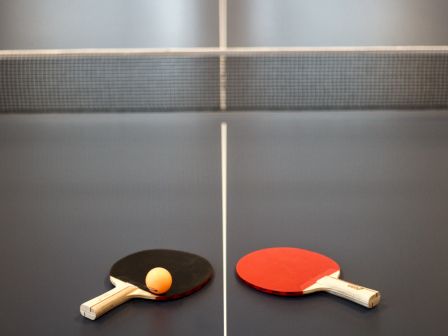 The image shows a table tennis setup with a net, two paddles (one black, one red), and a yellow ball placed on a blue table.