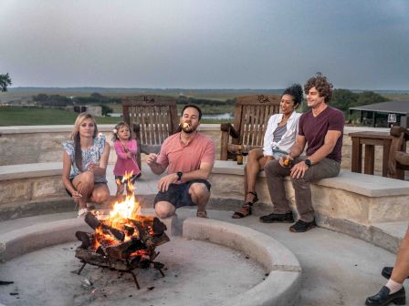 A group of people roasting marshmallows around a fire pit, enjoying a relaxed outdoor gathering in a scenic setting.