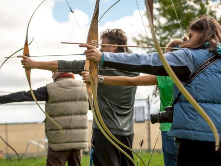 Multiple people are practicing archery outdoors with wooden bows, with one person wearing a blue jacket and camera.