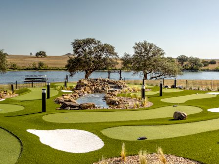 An outdoor mini golf course with artificial grass, a scenic pond, trees, and a body of water in the background, set under a clear blue sky.