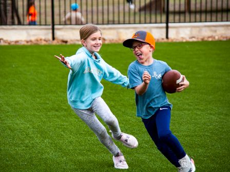 Two kids are playing on a grassy field, one holding a football and running while the other reaches out, both smiling and having fun.