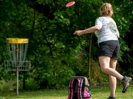 A person is playing disc golf, aiming a disc towards a metal basket on a grassy area, with a backpack on the ground nearby.