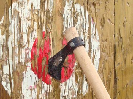 An axe is embedded in a wooden target with a red bullseye, indicating it has been used for axe throwing.