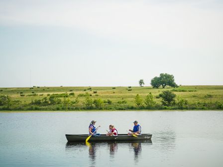 Three people are in a canoe on a calm body of water with a grassy landscape in the background.