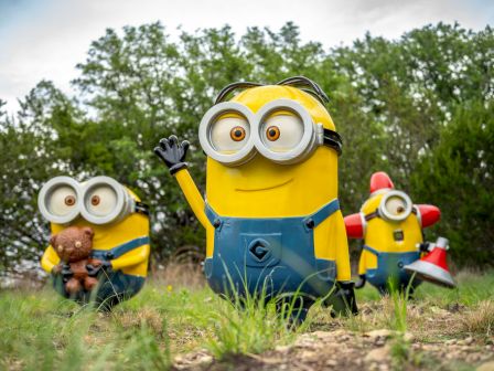 Several Minions in blue overalls playfully pose outdoors in a grassy forest clearing.