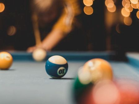 A close-up of a pool table showing several billiard balls, with a player blurred in the background preparing to take a shot.