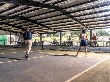 People playing pickleball indoors on a court with a net, under a metal roof, surrounded by trees in the background.