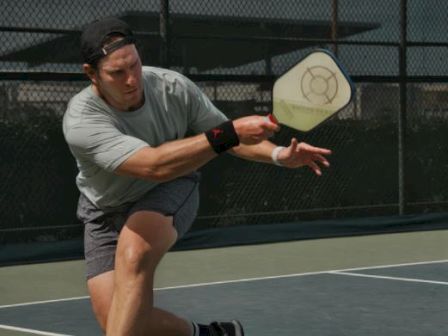 A person playing pickleball on an outdoor court, swung towards the ball with a paddle, wearing a cap, T-shirt, and shorts.