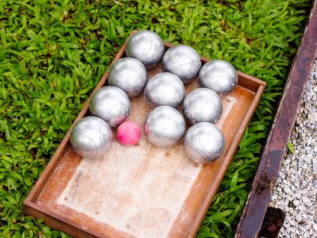 This image shows a wooden tray containing metal balls and a pink ball, placed on a grassy surface next to a gravel area.