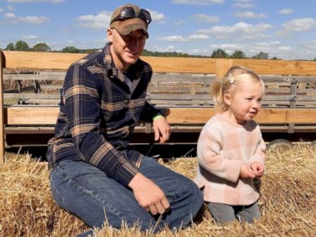 A man and a young girl are sitting on hay bales in a farm-like setting with a wooden fence and a sunny, partly cloudy sky in the background.