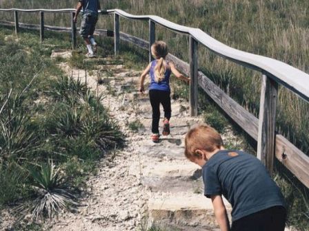 A man and two children are climbing a hill on a path with wooden railings on a sunny day. The path is surrounded by grassy terrain and rocks.