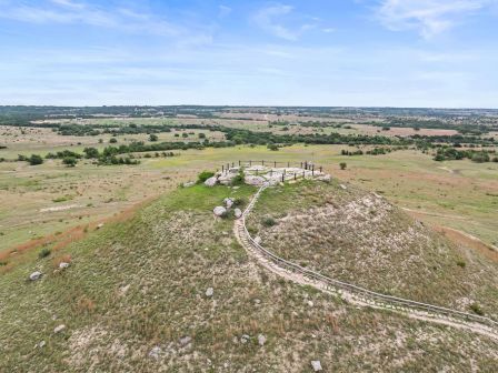 A drone view of a grassy hilltop with a fenced stone ruin and wooden posts overlooking open countryside.
