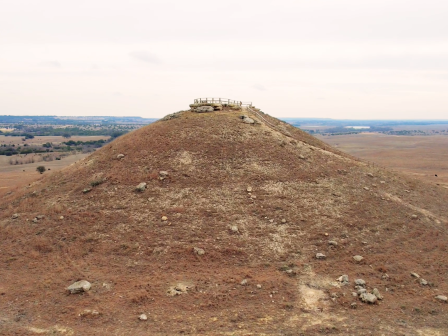A large, barren hill with a structure on top is surrounded by a vast, flat landscape and a winding dirt road leading towards it.