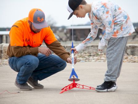 A person and a child are setting up a small rocket on a red launch pad outdoors, preparing for a launch together.