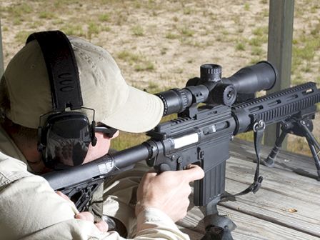 A person wearing a hat and ear protection is aiming through a rifle scope at an outdoor shooting range, lying on a wooden surface.