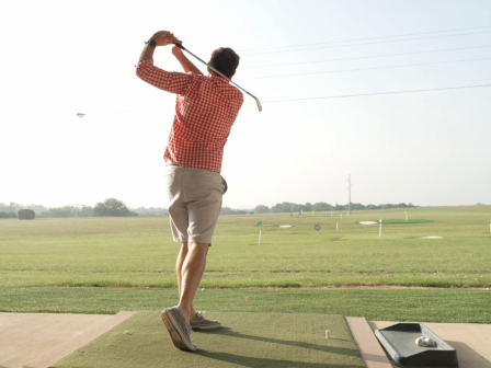 A person in a red checkered shirt swings a golf club at a driving range, with a grassy field in the background.