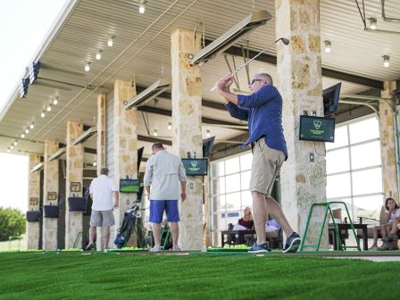 People are practicing their golf swings at an outdoor driving range with covered bays and seating areas.