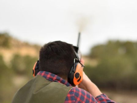 A person wearing ear protection and a checkered shirt is aiming and shooting a firearm outdoors, with smoke visible near the gun barrel.