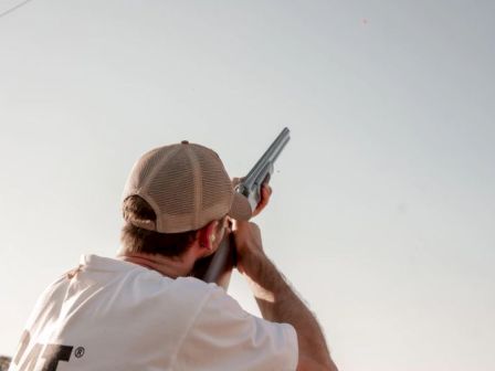 A man in a white shirt and beige cap is aiming a shotgun upward, possibly at a flying target, against a clear sky.