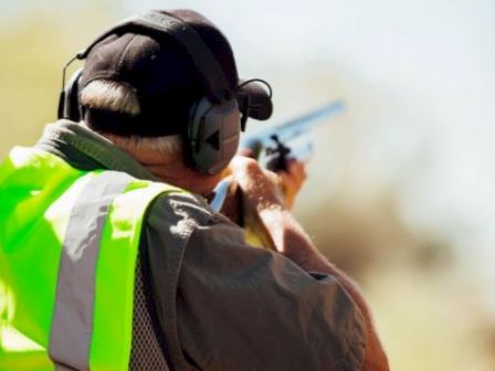 An individual in a high-visibility vest and protective gear aims a shotgun in an outdoor setting, suggesting they are engaged in shooting practice or sport.