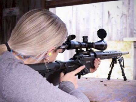 A person with blonde hair is aiming a scoped rifle at an outdoor shooting range while wearing safety glasses and ear protection.