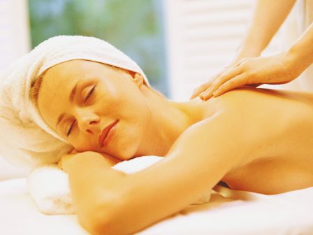 A woman relaxing on a massage table, eyes closed, receiving a shoulder massage with a towel wrapped around her head. Ending with a period.