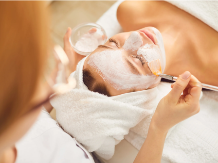 A person is applying a facial mask to a client lying down with a towel on their head in a spa setting.