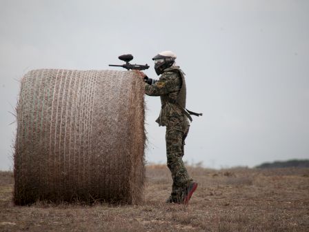 A person in camouflage with a paintball gun hides behind a large hay bale in an open field.