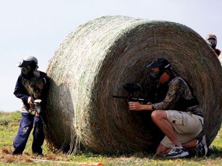 Two people in paintball gear hide behind a large hay bale while playing paintball outdoors in a grassy area.