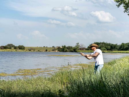 A man wearing a hat is fishing by a lake, surrounded by grass and trees under a partly cloudy sky.