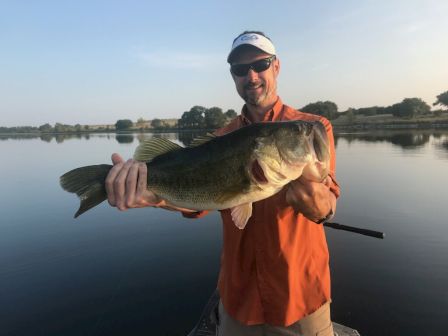 A person in an orange shirt and cap is holding a large fish by a lake, smiling under clear skies, with trees in the background and calm water.