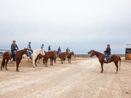 A group of people on horseback in a rustic outdoor setting, with a few structures in the background, under a cloudy sky.