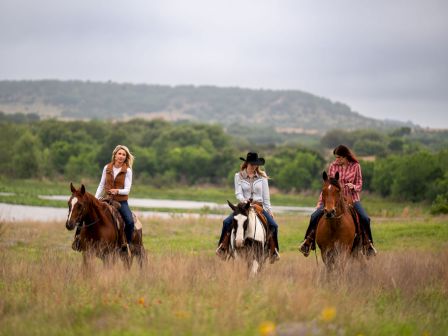 Three people horseback ride through a grassy countryside with hills in the background, enjoying an outdoor trail ride.