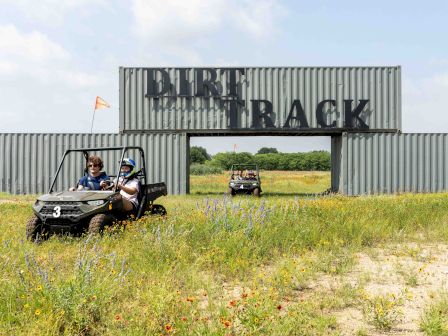 People driving buggies on a grassy field under a sign that reads "Dirt Track," with flowers and trees in the backdrop.