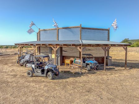 The image shows an outdoor pavilion with a metal roof, housing several off-road vehicles and two checkered flags on top.