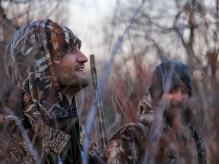 Two people in outdoor camouflage clothing standing among leafless trees and bushes, one looking upwards, possibly in a hunting scenario, under dim lighting.