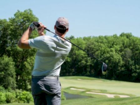 A person is swinging a golf club on a golf course, surrounded by lush greenery and trees, with a sand trap visible in the background.